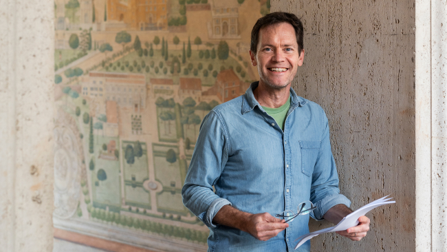 Color photo of a light skinned man in a blue button up shirt standing in front of a painted mural, smiling at the camera