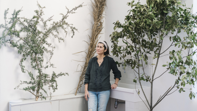 Color photo of a light skinned woman in an artist studio with three large plants or trees in the room