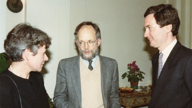 Color photograph of three light skinned people in a room talking to each other