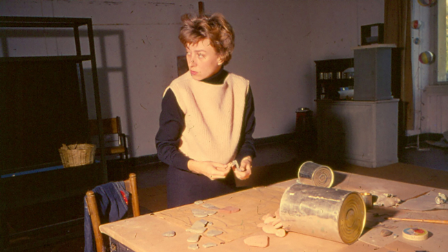 A color photo from the early 1960s depicting a light skinned woman in an artist's studio, studying a collection of small stones on a work table