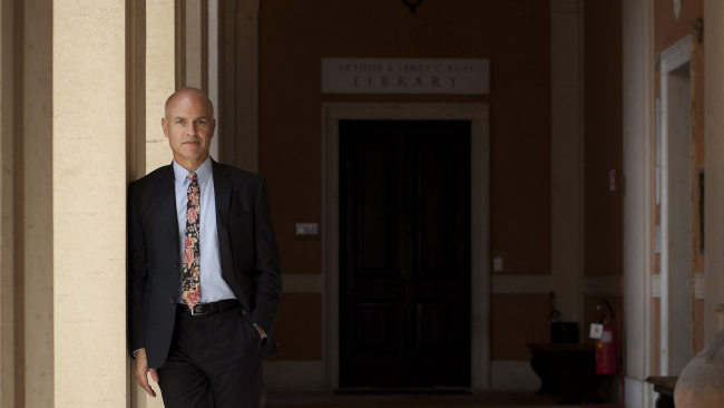 Color photograph of a light skinned man wearing a suit and tie and standing in the interior courtyard of a building in Rome