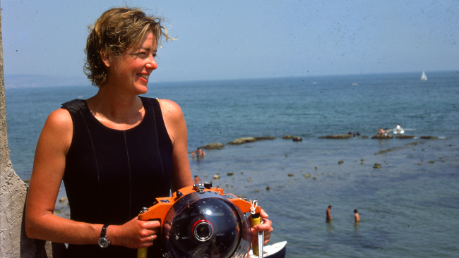 A woman in profile wearing a black wetsuit and holding an underwater camera, the sea behind her; she stares off into the distance