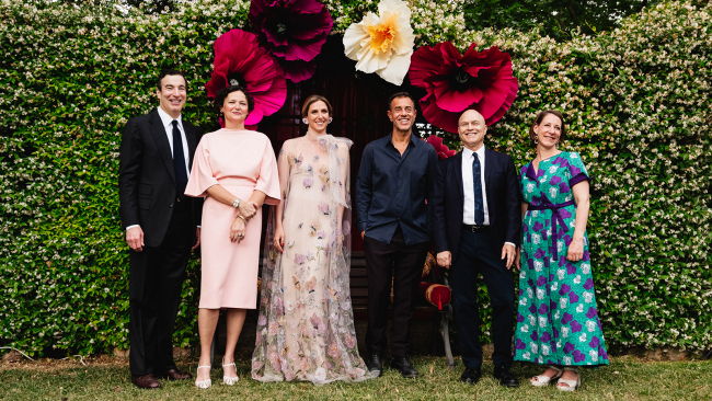 A group of six men and women wearing gala attire and posting in front of a green hedge with large artificial flowers above them