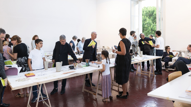 Color photo of an artist studio with many people in the room looking at objects on tables