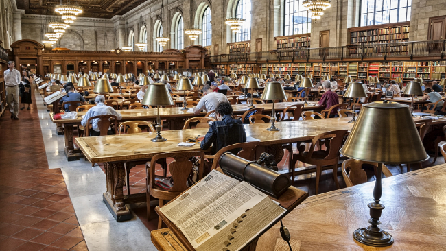 Color panoramic photograph of the Rose Reading Room at the New York Public Library, with an open dictionary on a stand in the foreground and rows of tables, lamps, chairs, and people going back into the far distance