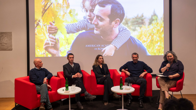 Color photo of five people sitting in red chairs on a stage in a lecture room, with a video projection of a child on a man's shoulders behind them