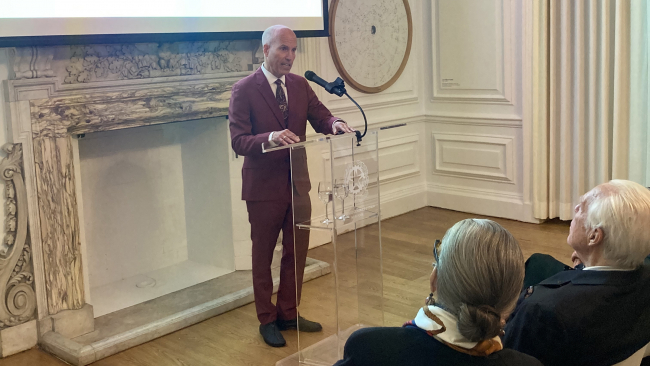 Color photograph of a light skinned man wearing a dark red suit speaking at a lecturn