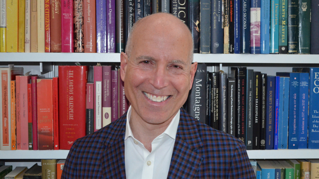 Color photograph of the head and shoulders of a light skinned man wearing a dark blazer and white shirt; he is positioned in front of a shelf of books with colorful spines as he smiles and looks at the camera