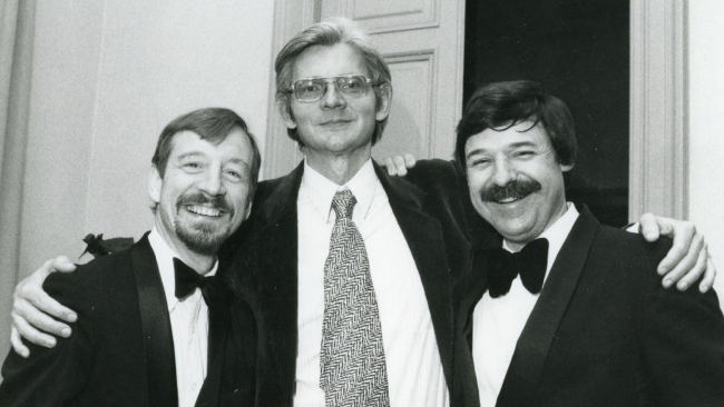 Black and white photograph of the heads and torsos of three light skinned men posing for the camera: a tall man in the center wears a jacket and tie; the two shorter men flanking him wear jackets and bowties and sport mustaches