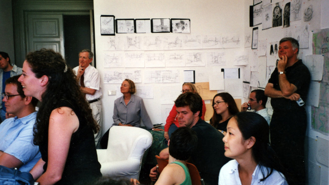 Color photograph of about a dozen people in an Academy studio, looking at an unseen person giving a presentation off camera; architectural drawings are pinned to the walls behind them